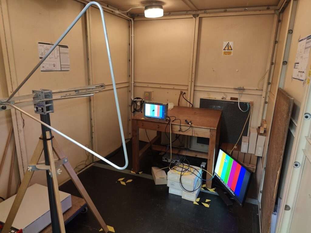 An EMC test chamber with a touchscreen unit on a wooden table and a 4K monitor on the floor, both showing colour bar test patterns, and a test antenna on a tripod in the foreground.