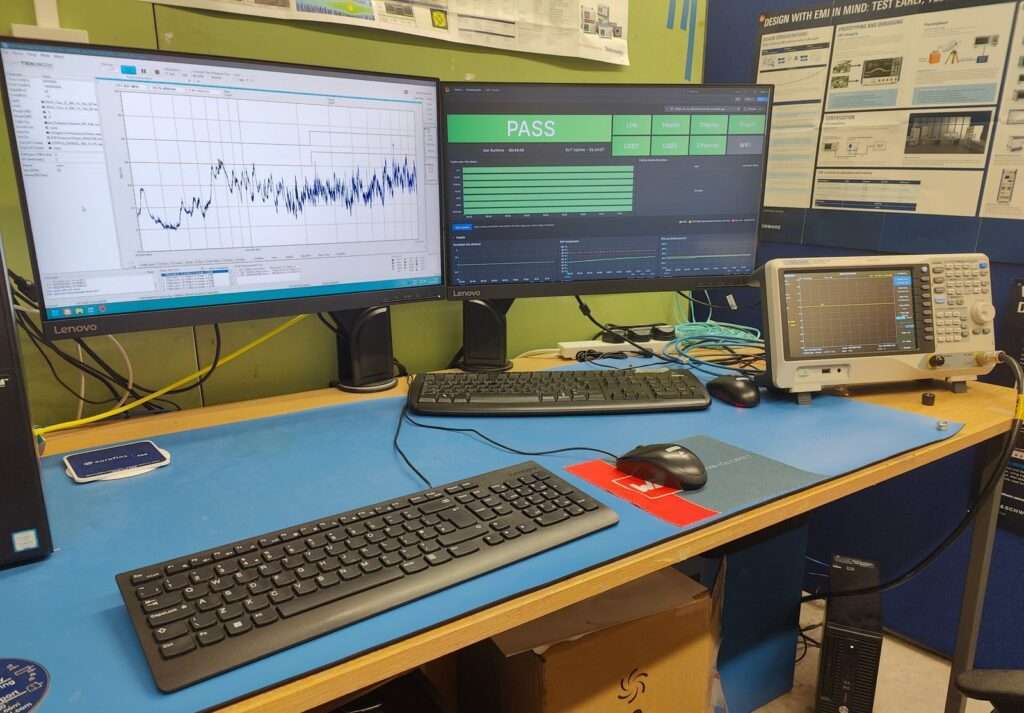 A test bench outside the EMC chamber with two monitors and a spectrum analyser. The right-hand monitor shows the EMC Health dashboard with a large green PASS tile.